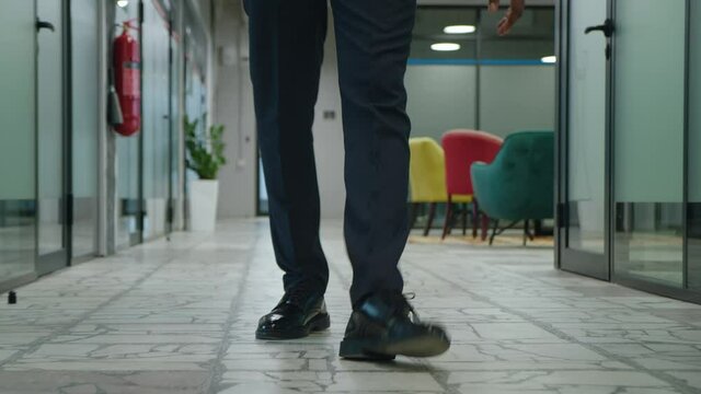 Front view feet of unrecognizable male entrepreneur walking through the open space office hallway. Tracking of legs of businessman in formal suit walking along office corridor.