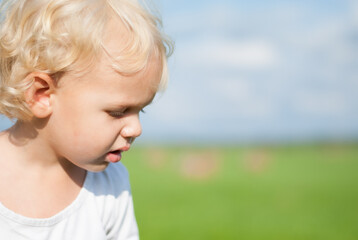 A small blonde curly-haired boy in a white T-shirt looks down and studies something against the background of blue sky and green grass