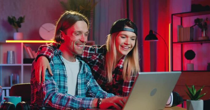 Lovely happy loving smiling blond girl hugging and kissing to the cheek her positive boyfriend when they together making purchases in online market on computer at home with night lighting