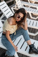 Fashionable beautiful girl with sunglasses in classic blue jeans, summer top blouse and boots sits on a beach lounger and looks at the camera
