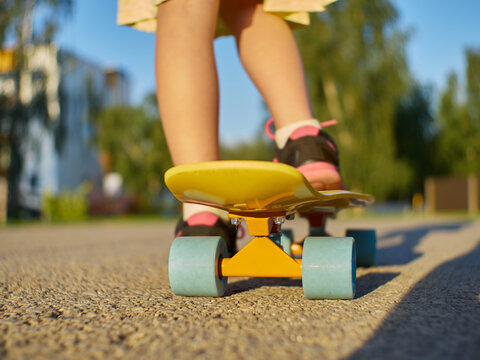 Girl kid standing on the asphalt road and yellow penny board with blue wheels on street background during evening time.
