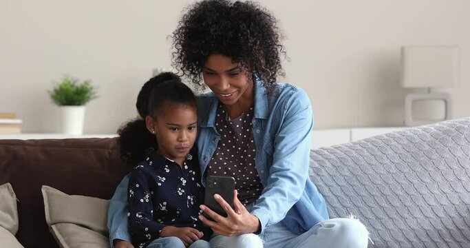 Smiling young african american woman showing funny mobile applications to cute little preschool child daughter, entertaining posing for selfie photo or playing online games together on smartphone.