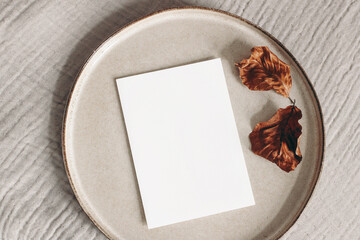 Naklejka na ściany i meble Autumn composition. Blank greeting card mockup and dry beech leaves on ceramic plate. Beige muslin table cloth textile background. Fall and Thanksgiving concept. Styled flat lay photography. Top view.