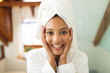 Portrait of smiling mixed race woman in bathroom, holding face after moisturising, looking at camera