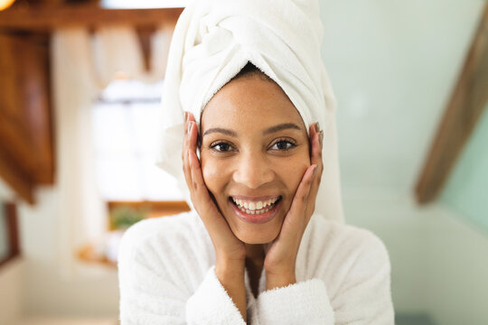 Fototapeta Portrait of smiling mixed race woman in bathroom, holding face after moisturising, looking at camera