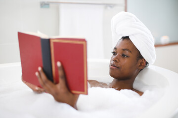 Happy african american woman in bathroom relaxing in bath reading book