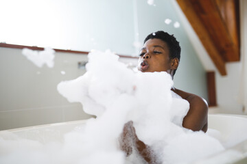 Happy african american woman relaxing in bath, having fun blowing bath foam