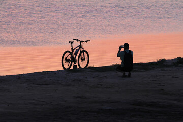 Man beside the bike takes a picture of the sunset near the water