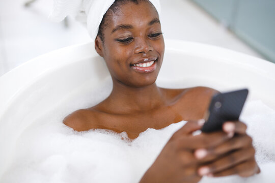 Smiling african american woman in bathroom, relaxing in bath using smartphone