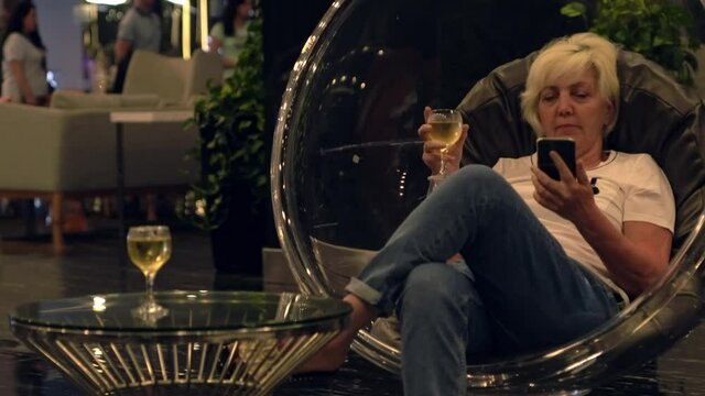 Woman relaxing in a hotel lobby in a modern perspex tub chair enjoying a glass of white wine and catching up on the news on her mobile phone