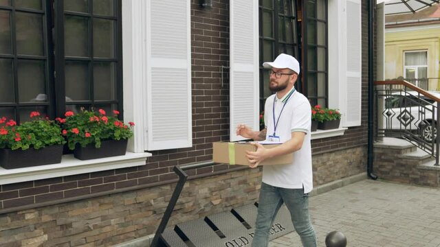 Young caucasian bearded postman with name tag, in white cap and glasses carrying box and going to door. Confident deliveryboy comparing house number with data in folder. Delivery concept.
