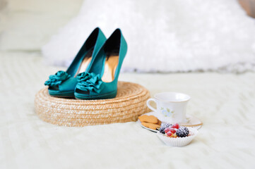 Morning still life. A small cake with fresh berries, a white porcelain teacup with cookies on a saucer and elegant turquoise-colored shoes with heels in the background in a blur.