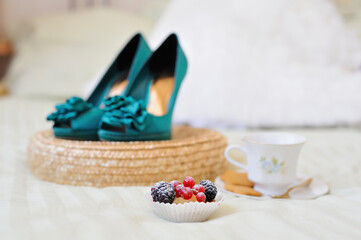 Obraz na płótnie Canvas Morning still life. A small cake with fresh berries, a white porcelain teacup with cookies on a saucer and elegant turquoise-colored shoes with heels in the background in a blur.