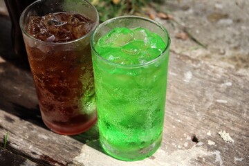 Cola and green soft drink with ice cubes on wood background