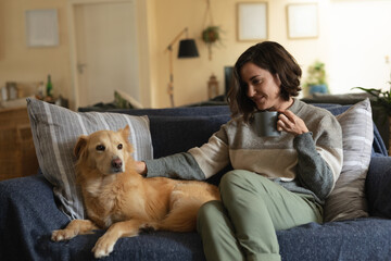 Smiling caucasian woman in living room sitting on sofa petting her pet dog and drinking coffee