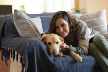Portrait of smiling caucasian woman in living room sitting on sofa embracing her pet dog