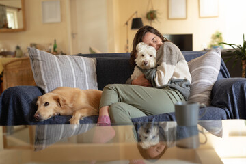 Smiling caucasian woman in living room sitting on sofa embracing her pet dog