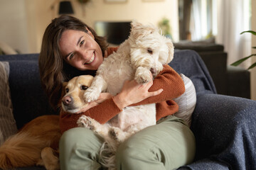Portrait of smiling caucasian woman in living room sitting on sofa embracing her pet dogs