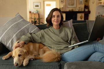 Portrait of smiling caucasian woman in living room, sitting on sofa with her pet dog, using laptop