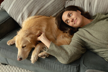 Smiling caucasian woman in living room, lying on sofa with her pet dog