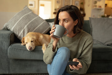 Caucasian woman in living room with her pet dog, using smartphone and drinking coffee