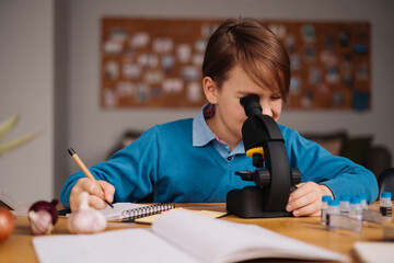 Naklejka na ściany i meble Distance online education, internet learning. First grade boy studying at home using microscope, making notes, biology online lesson