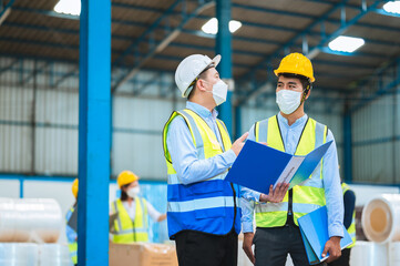 Team engineers and foreman wear a mask, hard hat, and vest. Standing, consult discuss industrial production management through data files with in mask factory. Employees working in the background.