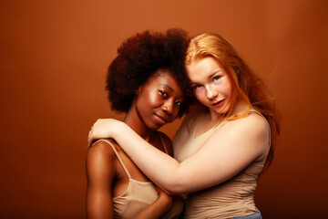 young pretty caucasian, afro woman posing cheerful together on brown background, lifestyle diverse nationality people concept