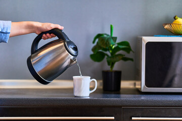 Female hands pouring boiling water from a modern metal stainless kettle in a glass cup for brewing tea in the kitchen at home