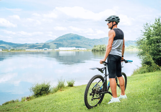 A middle-aged bicyclist man dressed in cycling clothes, helmet, and sunglasses with bicycle on grass hill enjoying lake and mountain landscape. Out-of-town sporty people activities concept image.