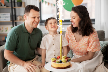 family, holidays and people concept - portrait of happy mother, father and little son with firework candle burning on birthday cake sitting on sofa at home party