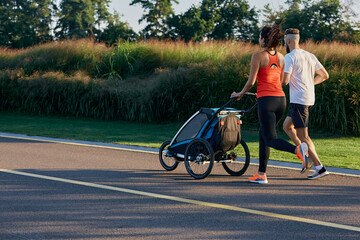Running young family with baby jogging stroller in motion enjoying workout and summer day in city...