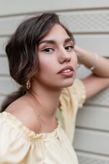 Outdoors portrait of pretty young woman with curly hairstyle in summer yellow fashion dress stands near vintage wall on the street and looks at the camera