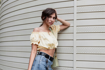 Pretty young girl in a yellow blouse with blue jeans and a fashionable handbag stands near the siding wall