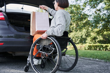 Fototapeta premium Girl sitting at the wheelchair is putting bags with purchases into car trunk