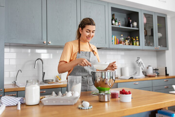 culinary, baking and people concept - happy smiling young woman cooking food on kitchen at home and sifting flour through sieve