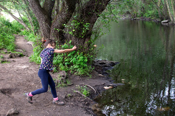 Naklejka na ściany i meble Camp girl throwing a stone into the river with her arm outstretched.