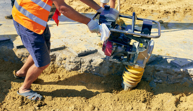 Closeup view of a person operating a vibratory earth rammer compacting the ground on the street, worker body parts are in background.