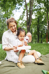 Full length portrait of happy young mother with cute little boy in park sitting on green grass together and enjoying Summer outdoors