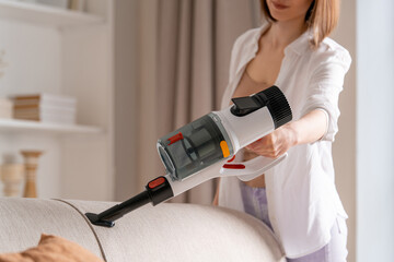 Young happy woman uses handheld wireless vacuum cleaner to clean the back of sofa. Close-up of hand...