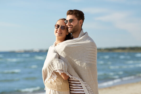 leisure, relationships and people concept - happy couple in sunglasses covered with blanket hugging on summer beach