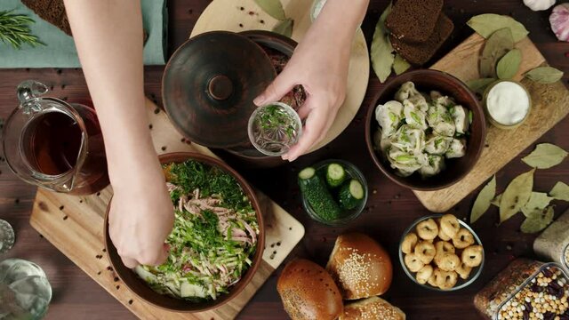 Traditional Russian Cuisine. Pouring kvass into salad with greenery, cooking okroshka, summer cold soup. Ukrainian food, borsch in bowl. Dumplings and sour cream, fresh homemade pastry top view.