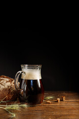 A glass jag with dark rye kvass or beer, with a thick foam. On a wooden board with crackers and spikelets. Rye round bread in the background