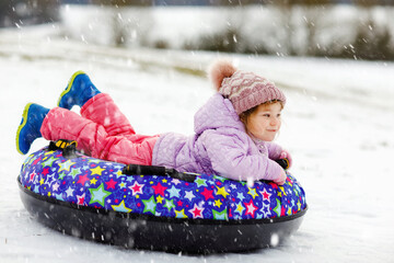 Active toddler girl sliding down the hill on snow tube. Cute little happy child having fun outdoors...