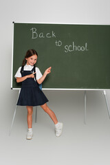 full length view of schoolgirl pointing at chalkboard with back to school lettering on grey