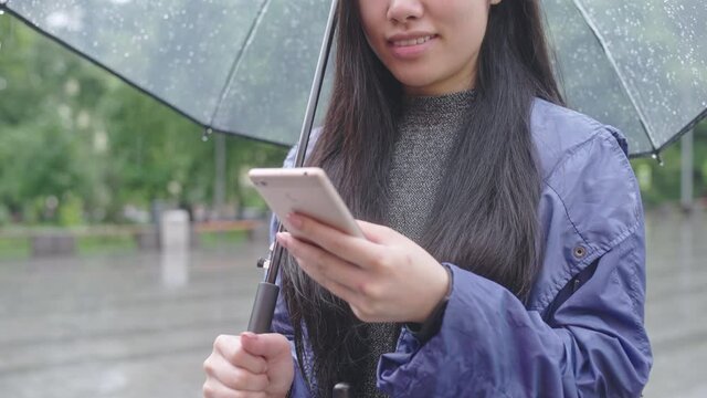 Young asian woman smiling, chatting with friend on smartphone, standing in rain