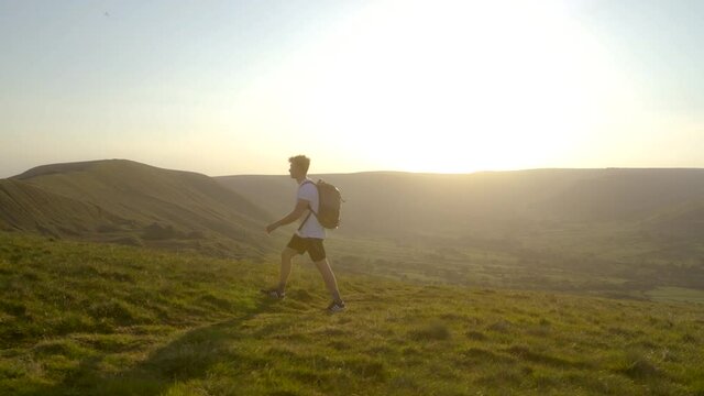 Stabilised gimbal shot of man with backpack walking on top of Mam Tor, Castleton, Peak District, England at sunset. View of sunset, valley, green fields, rolling hills and clear skies.
