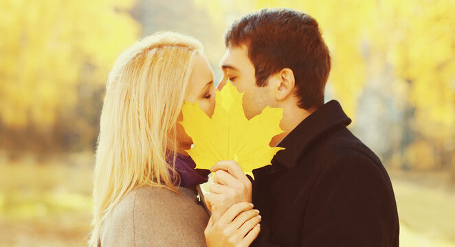 Portrait of happy young loving couple kissing covering her lips to yellow maple leaves in warm sunny autumn day