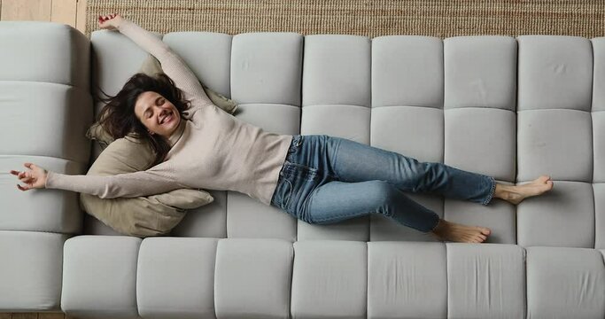 Top Above View Happy Young Beautiful Smiling Woman Falling On Huge Comfortable Sofa, Raising Arms Stretching Back Muscles, Enjoying Carefree Peaceful Weekend Relaxation Time Alone In Living Room.