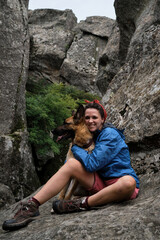 Female tourist on mountain hike in blue jacket and boots. Pretty young Caucasian woman is sitting on rock and hugging dog. Hiking in mountains with German Shepherd.